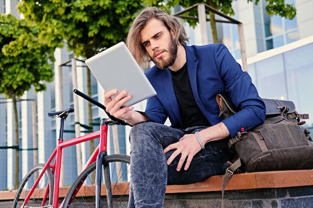 bärtiger Mann mit langen blonden Haaren hält Tablet-PC mit rotem Single-Speed-Fahrrad in einem Park im Hintergrund.