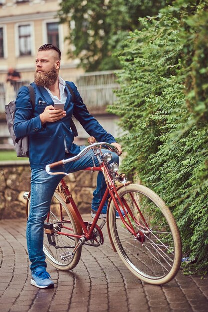 Bärtiger Mann mit stylischem Haarschnitt in Freizeitkleidung mit Rucksack, sitzend auf einem Retro-Fahrrad in einem Stadtpark.