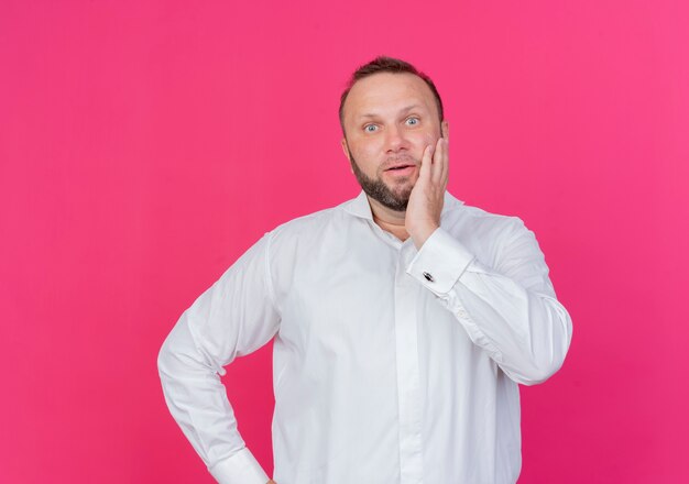 Bärtiger Mann mit weißem Hemd verwirrt über rosa Wand stehend