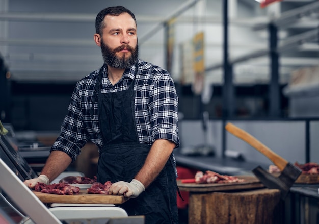 Bärtiger Metzger in einem Fleecehemd, der auf einem Markt frisch geschnittenes Fleisch serviert.