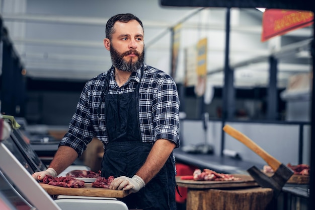 Bärtiger Metzger in einem Fleecehemd, der auf einem Markt frisch geschnittenes Fleisch serviert.