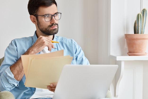 Bärtiger nachdenklicher Mann arbeitet von zu Hause aus, zählt Finanzdaten, hält Papierdokumente