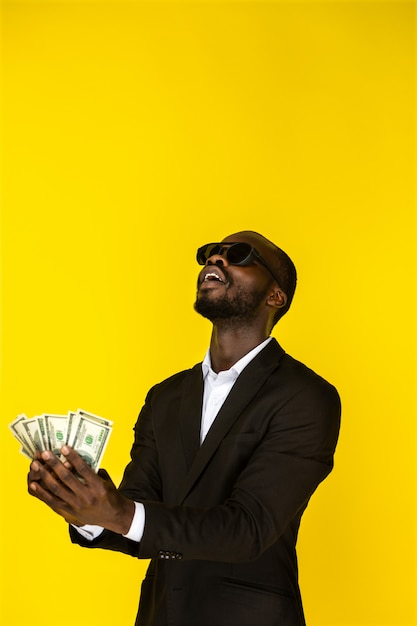 Bärtiger stilvoller junger afroamerikanischer Kerl hält Dollar in beiden Händen und wird sie oben werfen, Sonnenbrille und schwarzen Anzug tragend