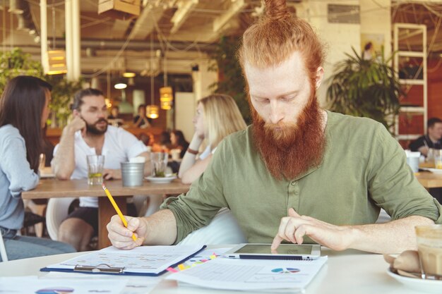 Bärtiger Student, der an Kurspapieren arbeitet, Nachforschungen anstellt, Statistiken auf Tablets analysiert und mit Bleistift in ein Notizbuch schreibt.