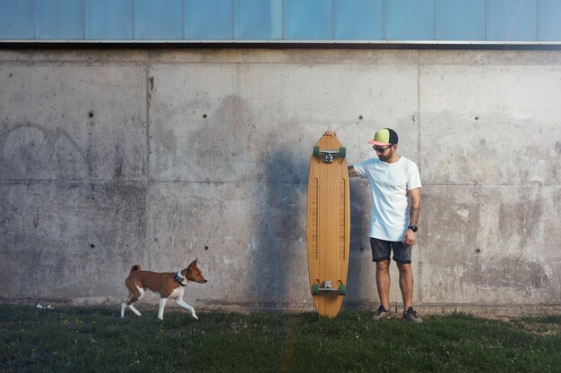 Bärtiger und tätowierter Longboarder, der neben einer Betonwand steht und einen sich nähernden braunen und weißen Basenji-Hund betrachtet