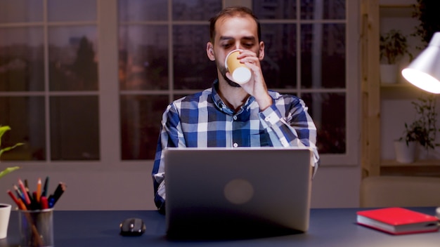 Bärtiger Unternehmer trinkt einen Schluck Kaffee und arbeitet nachts in seinem Heimbüro am Computer.