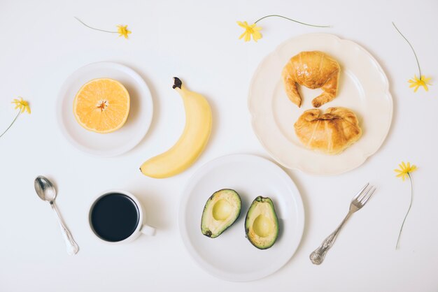 Banane; Croissants; halbierte Avocado; Kaffeetasse auf weißem Hintergrund mit Löffel und Gabel auf weißem Hintergrund