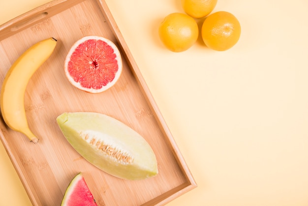Banane; Grapefruit; Wassermelone; und Muskmelon auf hölzernen Tablett nahe den ganzen Orangen gegen beige Hintergrund