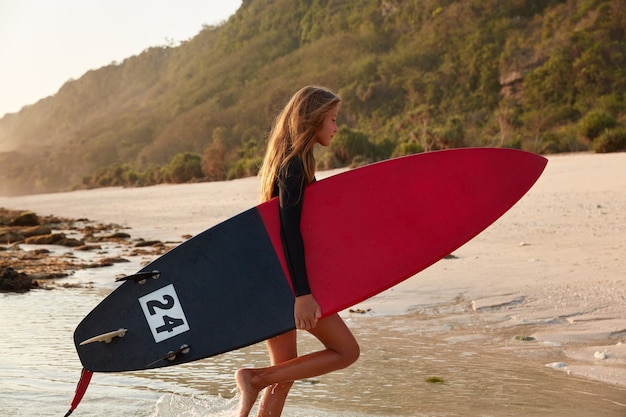 Barfuß Frau steht seitlich, hält Surfbrett, genießt Freizeit zum Surfen, posiert im Meer in Küstennähe, gegen Felsen