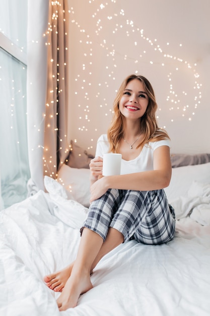 Barfuß glückliches mädchen, das mit tasse tee und lichtern an der wand aufwirft. innenaufnahme der entspannten europäischen blonden dame, die auf dem bett mit gekreuzten beinen sitzt.