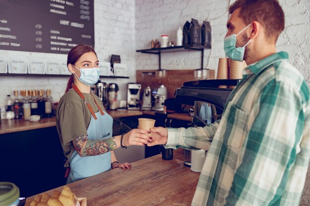 Barista in medizinischer maske, die dem kunden im café kaffee gibt