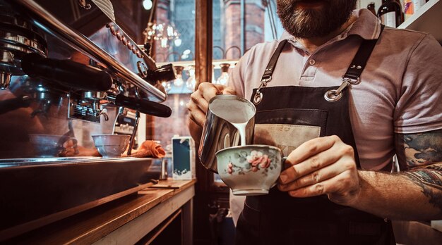 Barista in Schürze macht einen Cappuccino, gießt Milch in eine Tasse in einem Restaurant oder Café