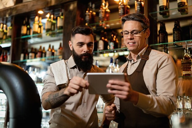 Barista mittleren Alters und sein junger Kollege verwenden Touchpad, während sie in einer Kneipe arbeiten