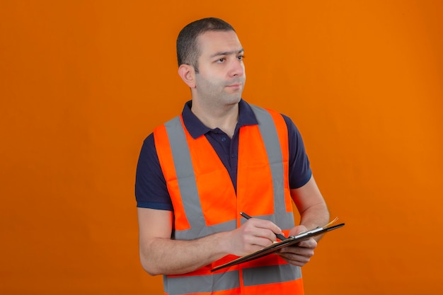 Bauarbeiter, der Weste mit einem ernsten Gesicht trägt, das weg schaut, um Zwischenablage mit Stift in Händen lokalisiert auf Orange zu halten