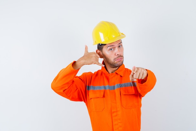 Bauarbeiter, der zeigendes Telefonzeichen in Uniform, Helm zeigt und zuversichtlich schaut. Vorderansicht.