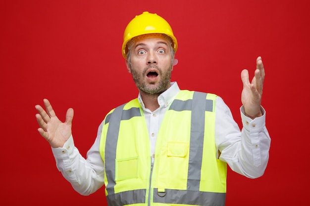 Bauarbeiter in Bauuniform und Schutzhelm, der erstaunt und überrascht in die Kamera blickt und die Arme über rotem Hintergrund hebt