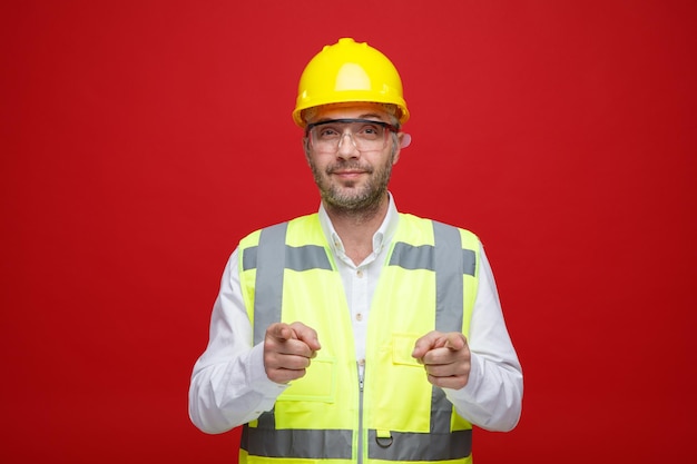 Bauarbeiter in Bauuniform und Schutzhelm mit Schutzbrille, der glücklich und positiv in die Kamera blickt und mit dem Zeigefinger auf die Kamera zeigt, die über rosa Hintergrund steht