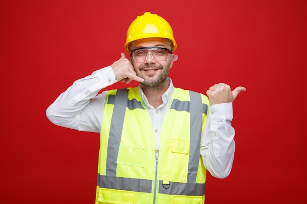 Bauarbeiter in Bauuniform und Schutzhelm mit Schutzbrille, der in die Kamera schaut, freundlich lächelt und mich anruft, Geste, die mit dem Daumen auf die Seite zeigt, die über rotem Hintergrund steht