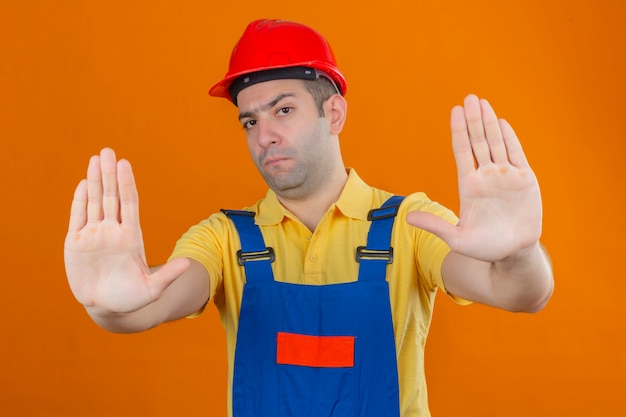 Kostenloses Foto bauarbeiter in uniform und rotem schutzhelm mit ernstem gesicht, das stoppschild lokalisiert auf orange lokalisiert