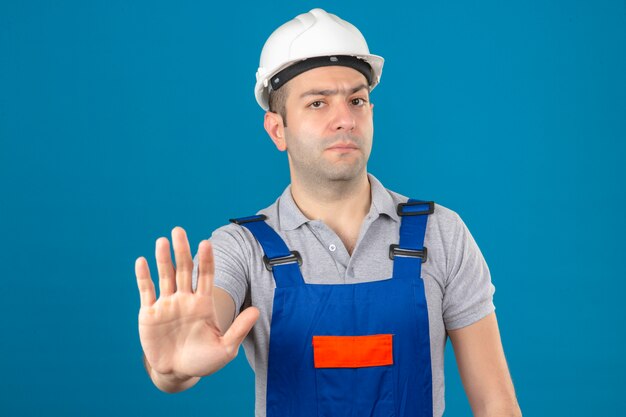 Bauarbeiter in Uniform und weißem Schutzhelm, der Stoppgeste mit Hand lokalisiert auf Blau macht