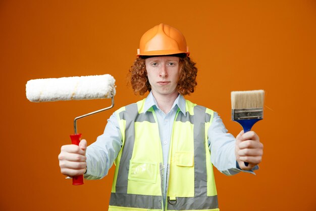 Bauarbeiter junger Mann in Bauuniform und Schutzhelm mit Farbroller und Pinsel, der mit ernstem Gesicht auf orangefarbenem Hintergrund in die Kamera blickt