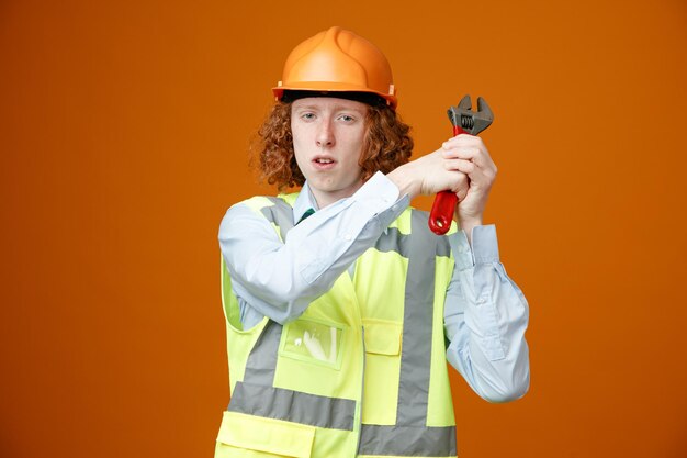 Bauarbeiter junger Mann in Bauuniform und Schutzhelm mit Schraubenschlüssel, der mit ernstem Gesicht auf orangefarbenem Hintergrund in die Kamera blickt