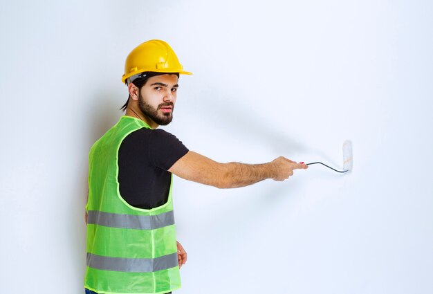 Bauarbeiter mit gelbem Helm, der die weiße Wand mit einer Trimmwalze malt.