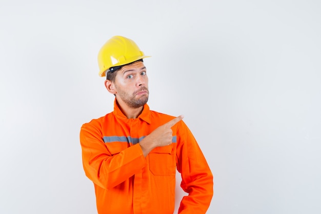 Bauarbeiter zeigt in Uniform, Helm und zögernd, Vorderansicht.