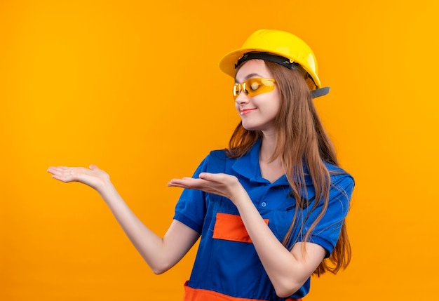 Bauarbeiterin der jungen Frau in der Bauuniform und im Sicherheitshelm, die beiseite schauen mit Lächeln auf Gesicht, das mit den Armen der Hände stehend darstellt