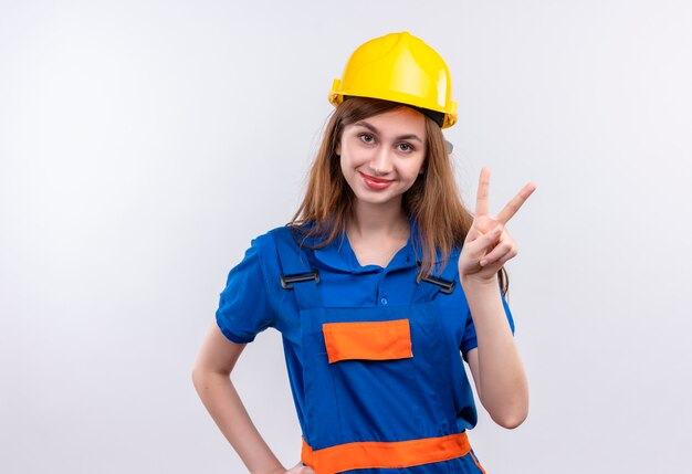 Bauarbeiterin der jungen Frau in der Bauuniform und im Sicherheitshelm, die das Siegeszeichen lächelnd freundlich stehend über weißer Wand zeigen