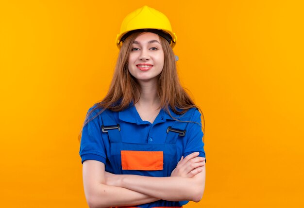 Bauarbeiterin der jungen Frau in der Bauuniform und im Sicherheitshelm, die mit verschränkten Armen stehen und mit dem selbstbewussten Lächeln auf Gesicht über orange Wand schauen
