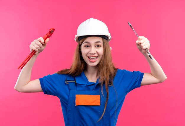 Bauarbeiterin der jungen Frau in der Bauuniform und im Sicherheitshelm, die Schraubenschlüssel in den erhöhten Armen halten, die breit lächelnd über rosa Wand lächeln