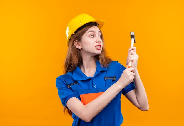 Bauherrin der jungen Frau in der Bauuniform und in der Sicherheitshelmschaukel, die über orange Wand steht