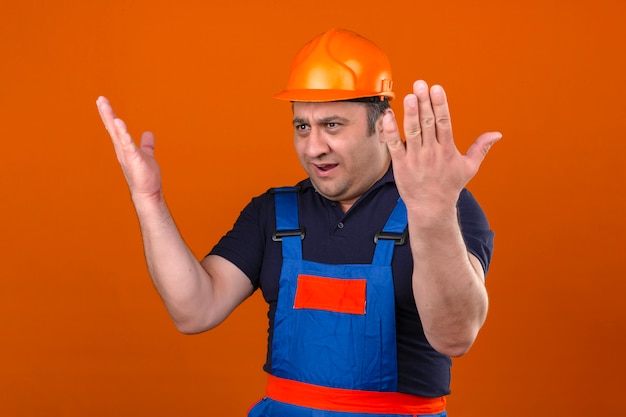 Baumeister, der Bauuniform und Schutzhelm trägt, hebt Arme mit Empörung über isolierte orange Wand