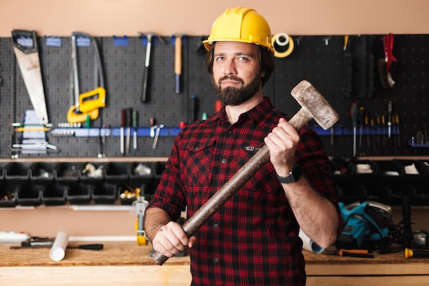 Kostenloses Foto baumeister in gelbem hardhat, der hammer in der hand hält und glücklich in die kamera schaut, mit werkzeugständer auf dem hintergrund in der werkstatt