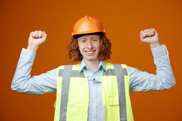 Kostenloses Foto baumeister junger mann in bauuniform und schutzhelm, der glücklich und aufgeregt in die kamera blickt und geballte fäuste hebt, die sich freuen, über orangefarbenem hintergrund zu stehen