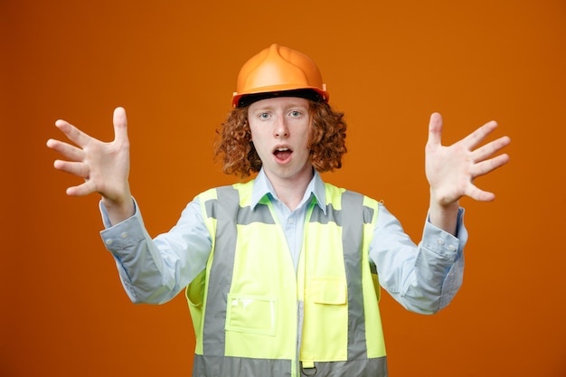 Baumeister junger Mann in Bauuniform und Schutzhelm, der in die Kamera schaut, verwirrt und überrascht ist, wie er die Arme hebt, die über orangefarbenem Hintergrund stehen