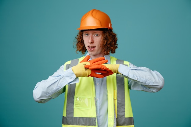 Baumeister junger Mann in Bauuniform und Schutzhelm mit Gummihandschuhen, der mit ernstem, selbstbewusstem Ausdruck in die Kamera blickt und mit Händen gestikuliert, die über blauem Hintergrund stehen