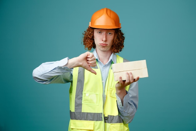 Baumeister junger Mann in Bauuniform und Schutzhelm mit zwei Ziegeln, die unzufrieden in die Kamera schauen und den Daumen nach unten zeigen, der über blauem Hintergrund steht