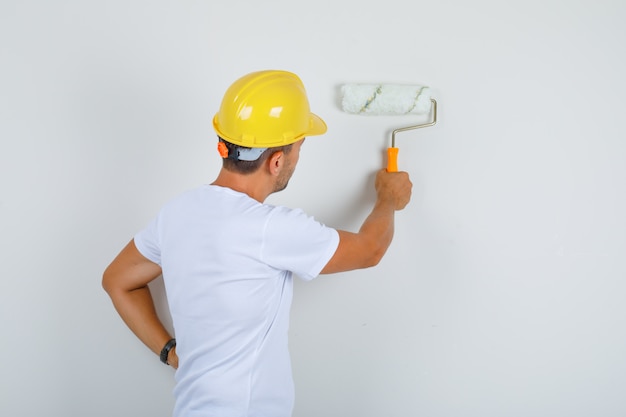 Baumeister malen Wand mit Rolle in weißem T-Shirt, Helm und suchen beschäftigt, Rückansicht.