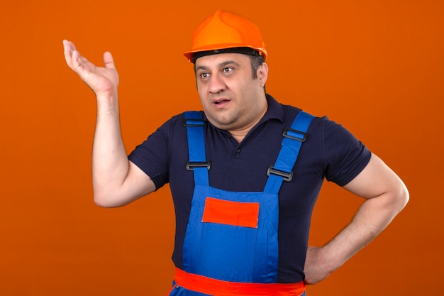 Baumeister Mann, der Bauuniform und Schutzhelm trägt, der mit erhobener Hand steht, ohne zu verstehen, was ahnungslos und verwirrter Ausdruck über isolierter orange Wand geschah