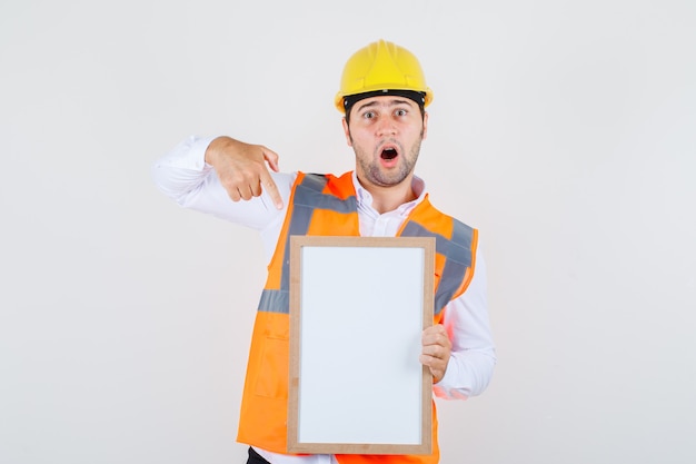 Baumeister Mann im Hemd, Uniform zeigt Finger auf weiße Tafel und schaut überrascht, Vorderansicht.