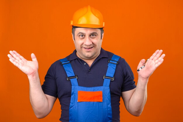 Baumeister Mann trägt Bauuniform und Schutzhelm zuckt mit den Schultern und spreizt die Hände, ohne zu verstehen, was ahnungslos und verwirrt über dem isolierten orangefarbenen Rücken passiert ist