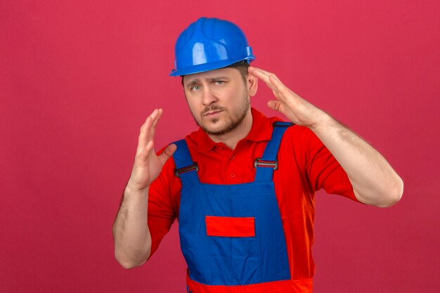 Baumeister Mann trägt Bauuniform und Sicherheitshelm missfallen und ängstlich stehend mit Händen über isolierte rosa Wand erhoben