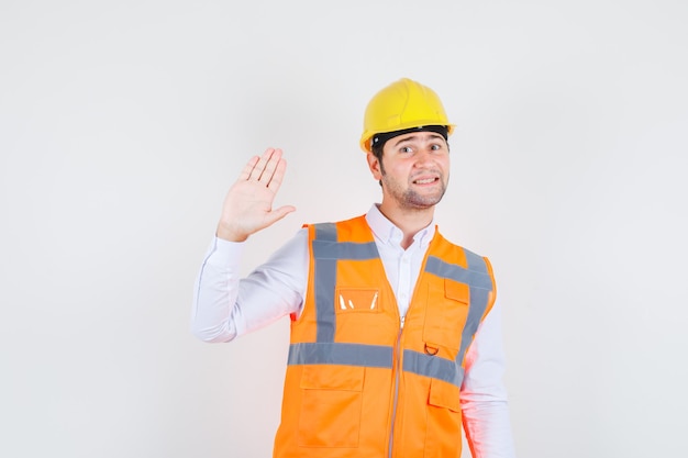 Baumeister Mann winkt Hand, um Hallo oder Auf Wiedersehen in Hemd, Uniform und lustig aussehen zu sagen. Vorderansicht.