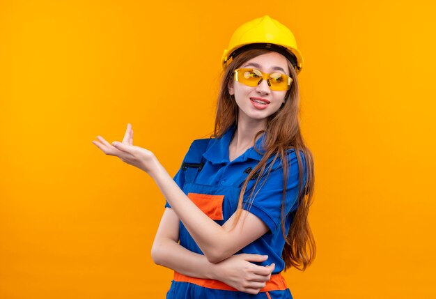 Baumeisterin der jungen Frau in der Bauuniform und im Sicherheitshelm, die mit erhabener Hand stehen, die mit skeptischem Ausdruck steht, der über orange Wand steht