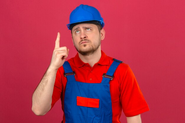 Baumeistermann, der Bauuniform und Sicherheitshelm trägt, der oben zeigt, zeigt mit dem Finger über isolierte rosa Wand