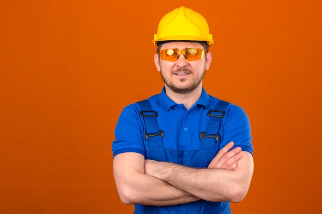 Baumeistermann, der eine einheitliche Baubrille und einen Sicherheitshelm trägt, die mit den Armen gekreuzt mit dem selbstbewussten Lächeln über der isolierten orange Wand stehen