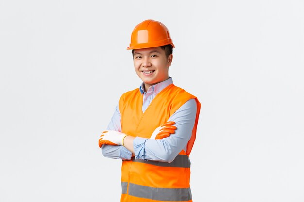 Bausektor und Industriearbeiterkonzept. Selbstbewusster junger asiatischer Ingenieur, Bauleiter in reflektierenden Kleidern und Helm, verschränkten Armen und frech lächelnd, für Qualität, weiße Wand