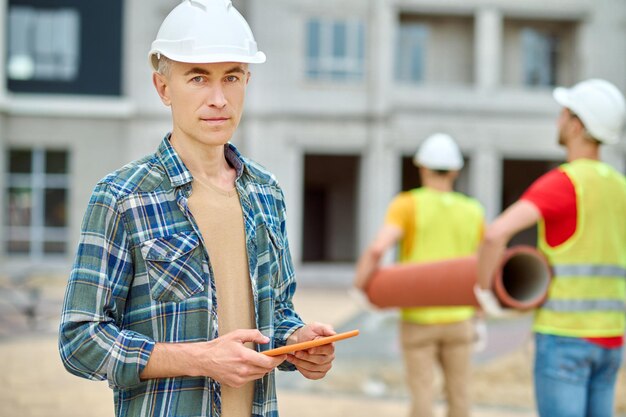 Baustelle. Nachdenklicher Mann in Schutzhelm und kariertem Hemd mit Tablet, das in die Kamera schaut und Arbeiter, die auf der Baustelle ein Rohr hinter sich tragen